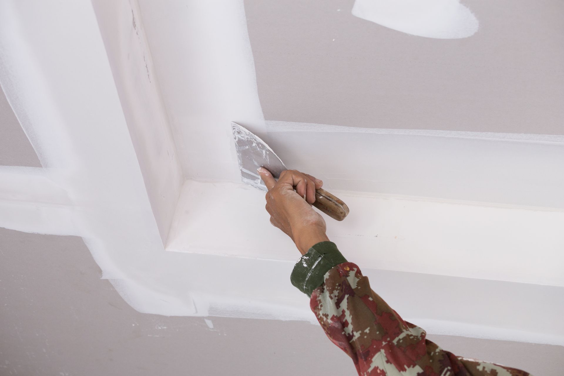 Person using a putty knife to smooth white drywall ceiling seams.