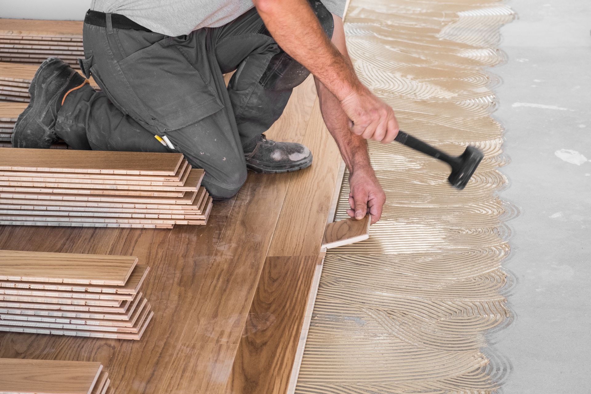 Worker spreading wood floor adhesive with a trowel beside stacked flooring planks