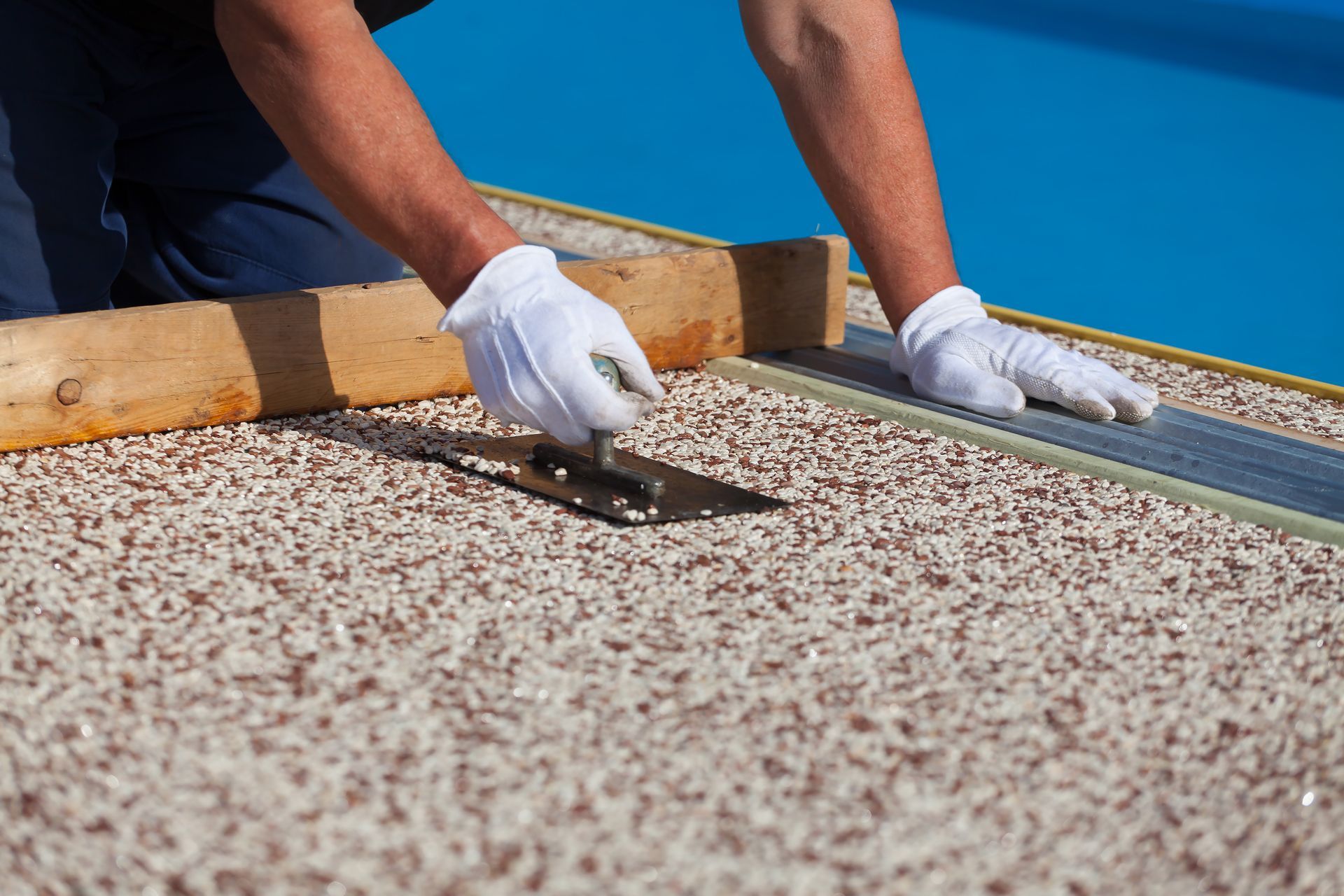 Worker spreading roofing material on a flat roof with a trowel, wearing white gloves.