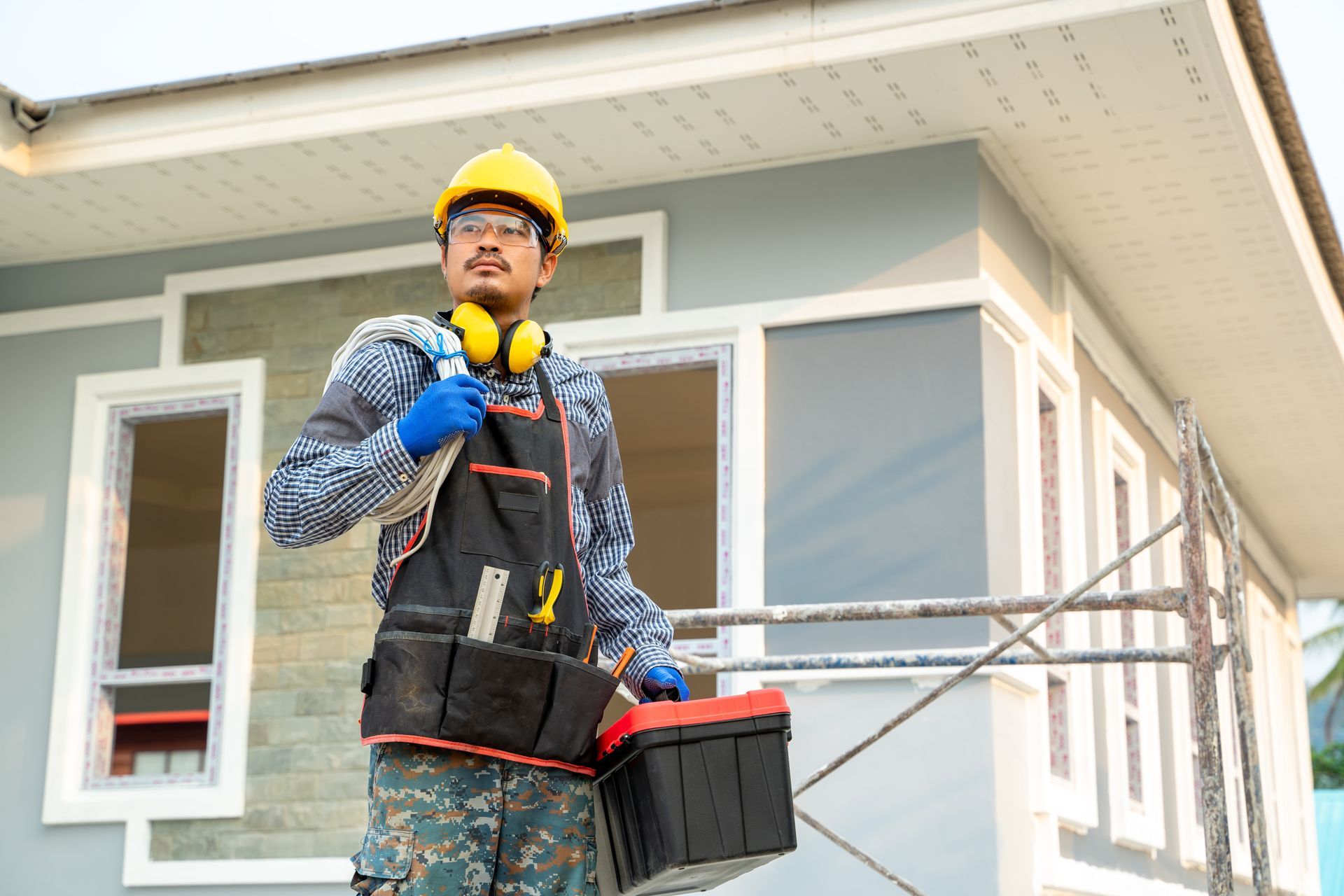 Construction worker in a hard hat and gloves standing outside a house, holding tools and a toolbox.
