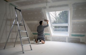 Person kneeling and painting drywall in a room with a ladder and window