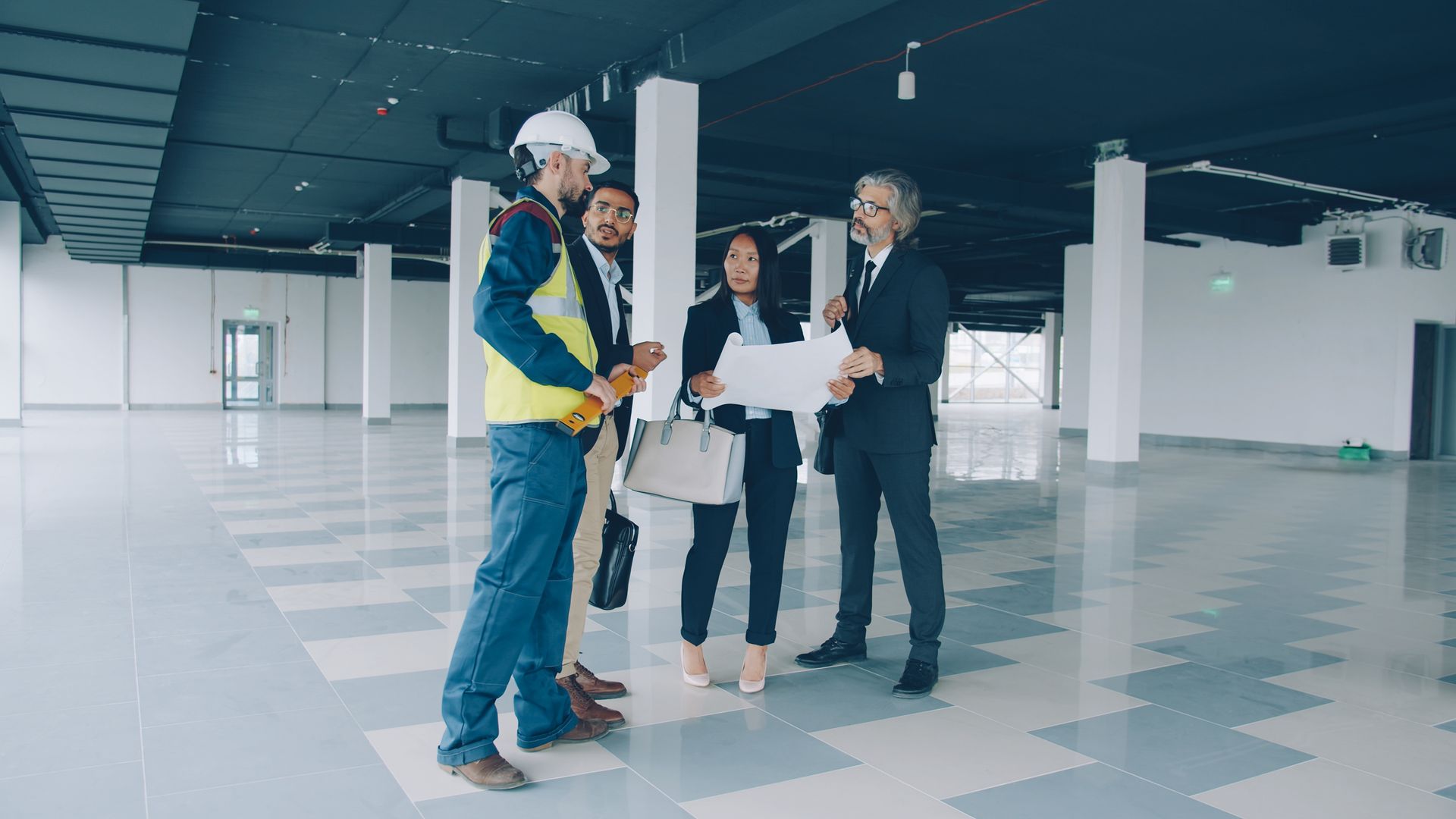 Four people in hard hats discuss blueprints in an unfinished office space with tiled floors.