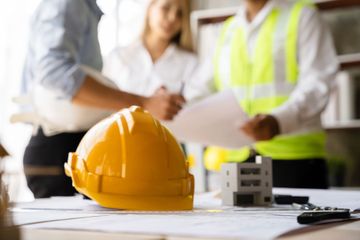 Yellow hard hat on blueprints at a construction planning meeting with people in the background