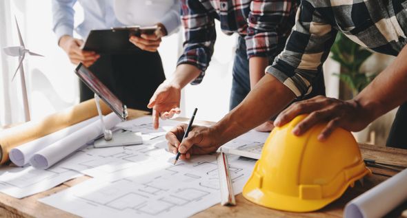 People collaborating over architectural plans beside a yellow hard hat on a desk