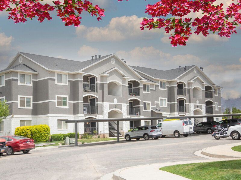 Apartment complex with gray siding, covered parking, and cars under pink blossoms on a sunny day