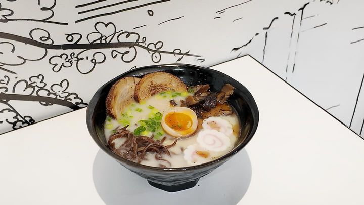 A bowl of ramen with sliced pork, boiled egg, and greens on a white table.