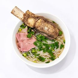 Bowl of noodle soup with beef slices, herbs, and a large braised bone on top
