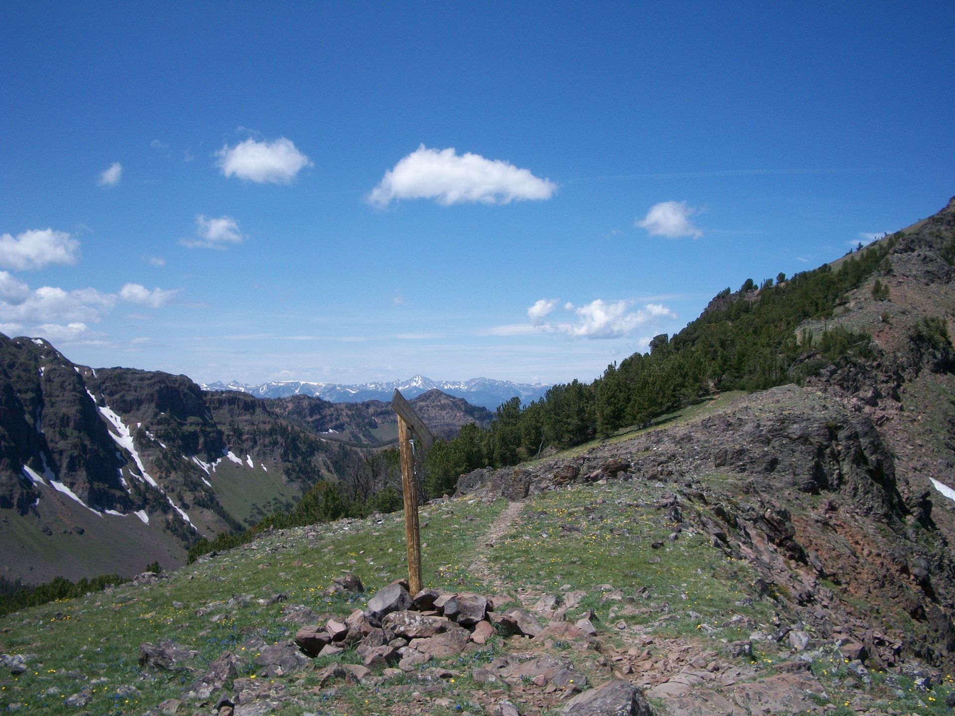Mountain trail overlook with a wooden post, rocky ground, green slopes, and distant snowy peaks under a blue sky.