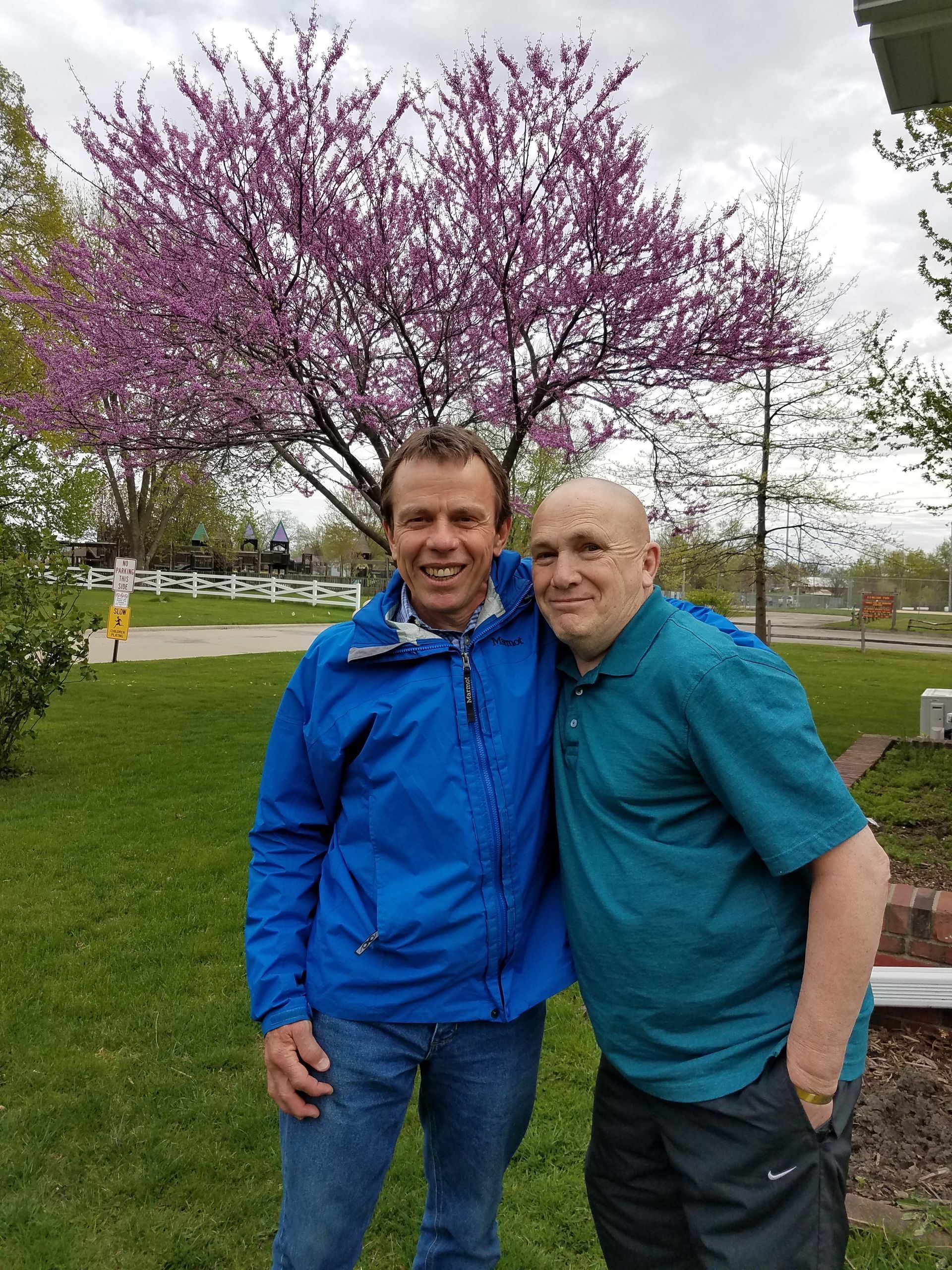 Two men standing together in a park beneath a blooming pink tree
