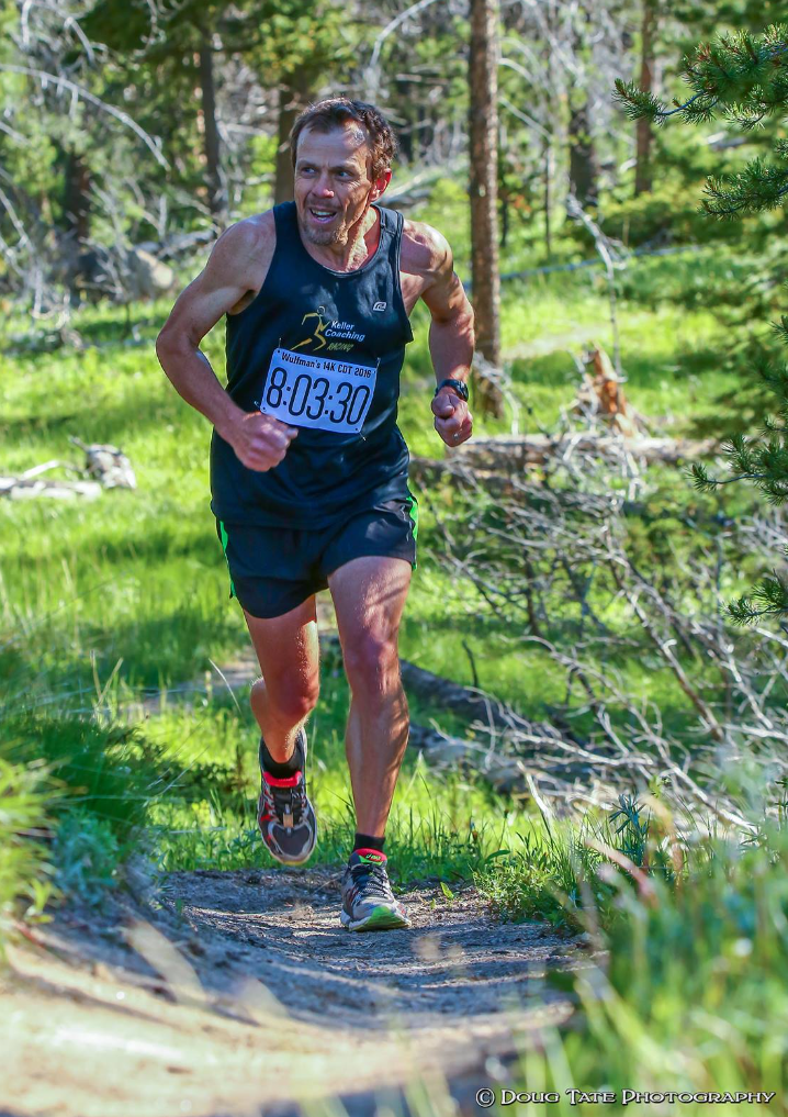 Runner in black gear with blue bib runs on a sunlit forest trail.
