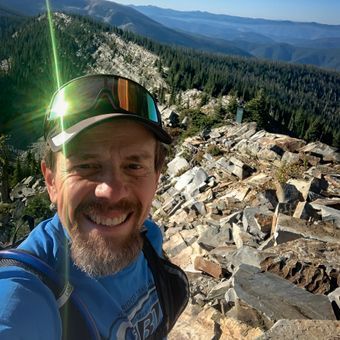 Smiling hiker taking a selfie on a rocky mountain summit with forested peaks in the background