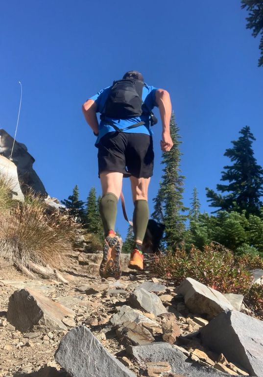 Hiker in blue shorts and backpack ascending a rocky mountain trail under a clear blue sky