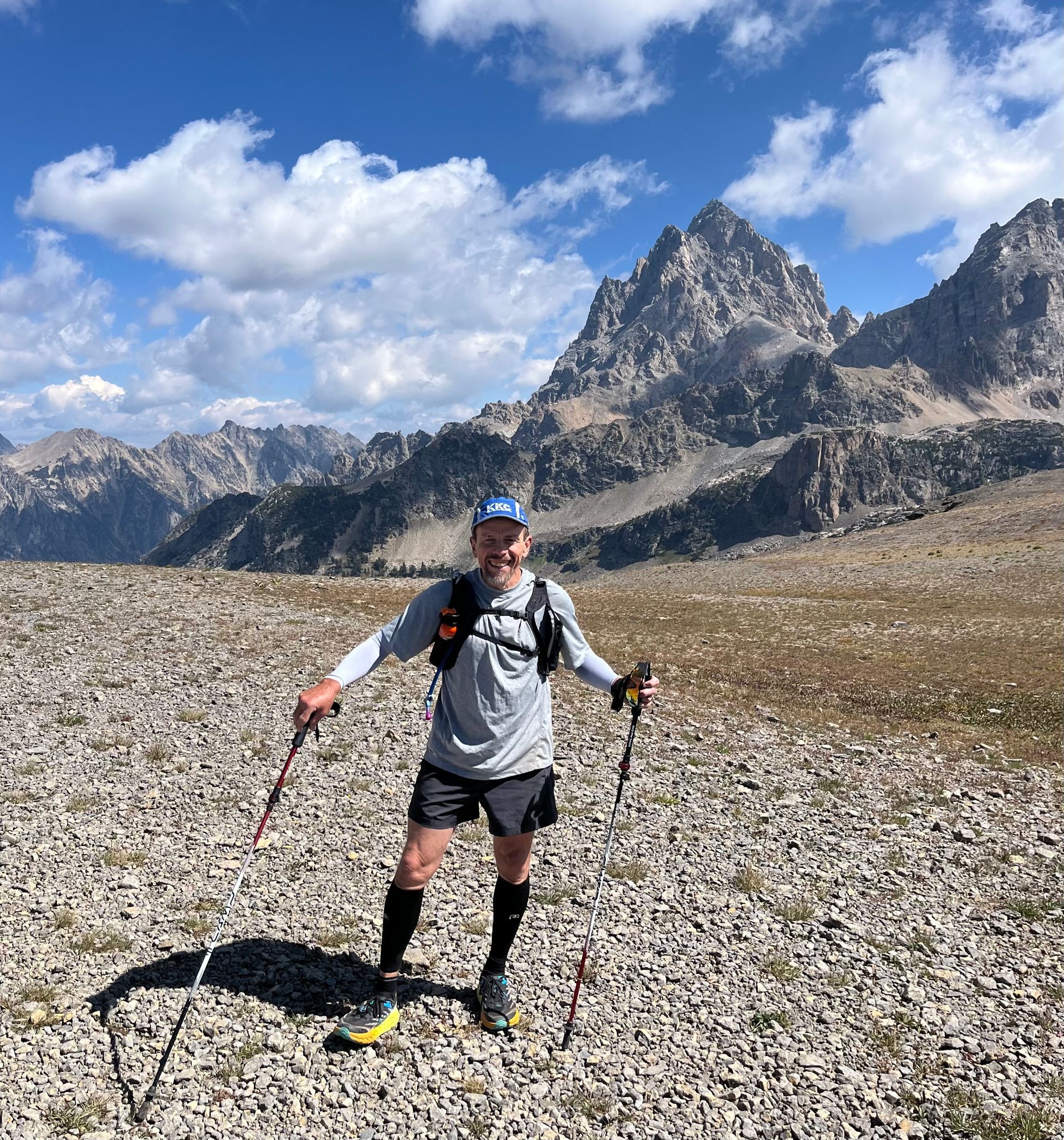 Hiker with trekking poles standing on a rocky alpine trail under a blue sky with mountains behind.