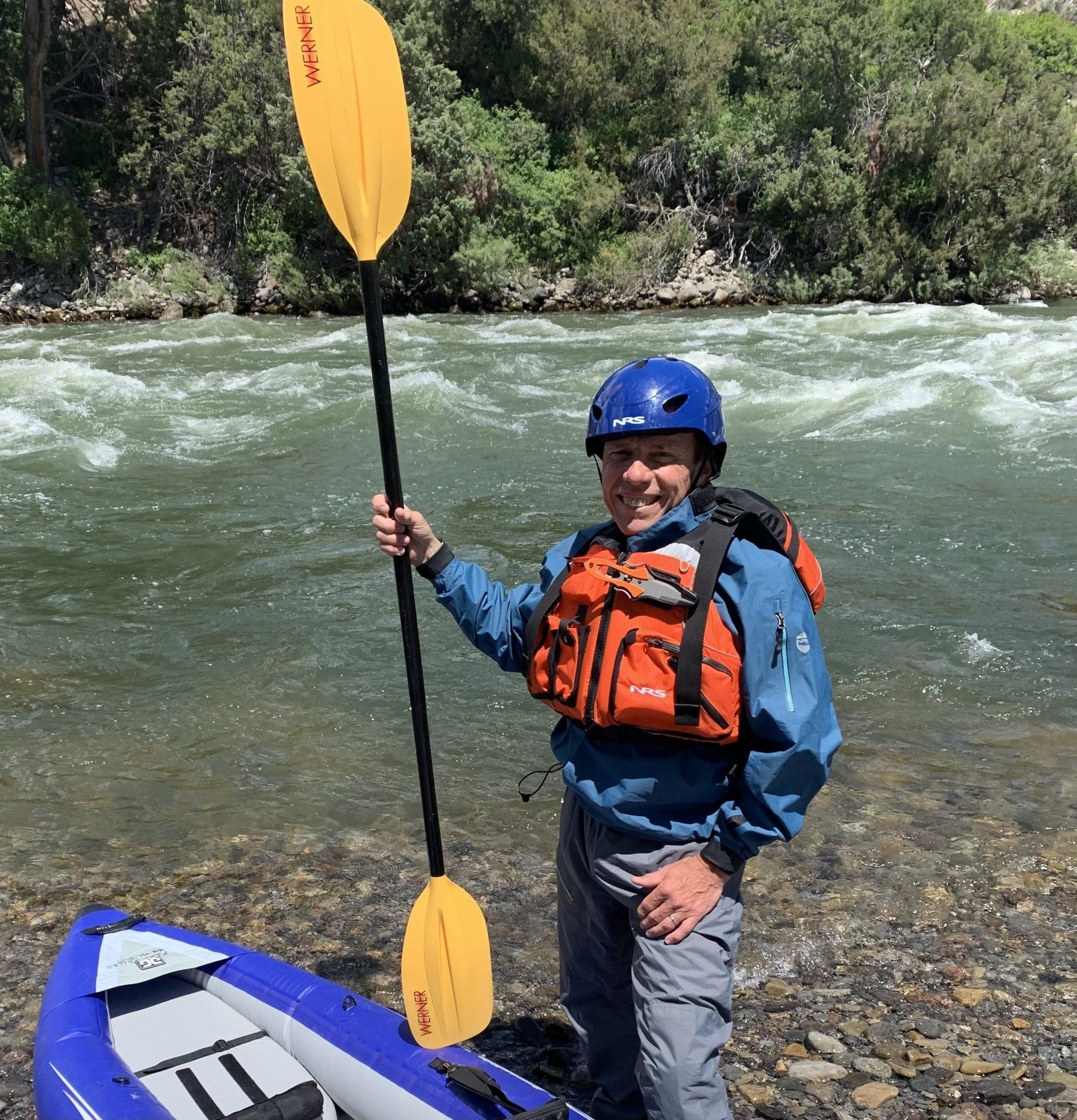 Smiling kayaker in blue helmet and orange vest holding a paddle beside a fast river.
