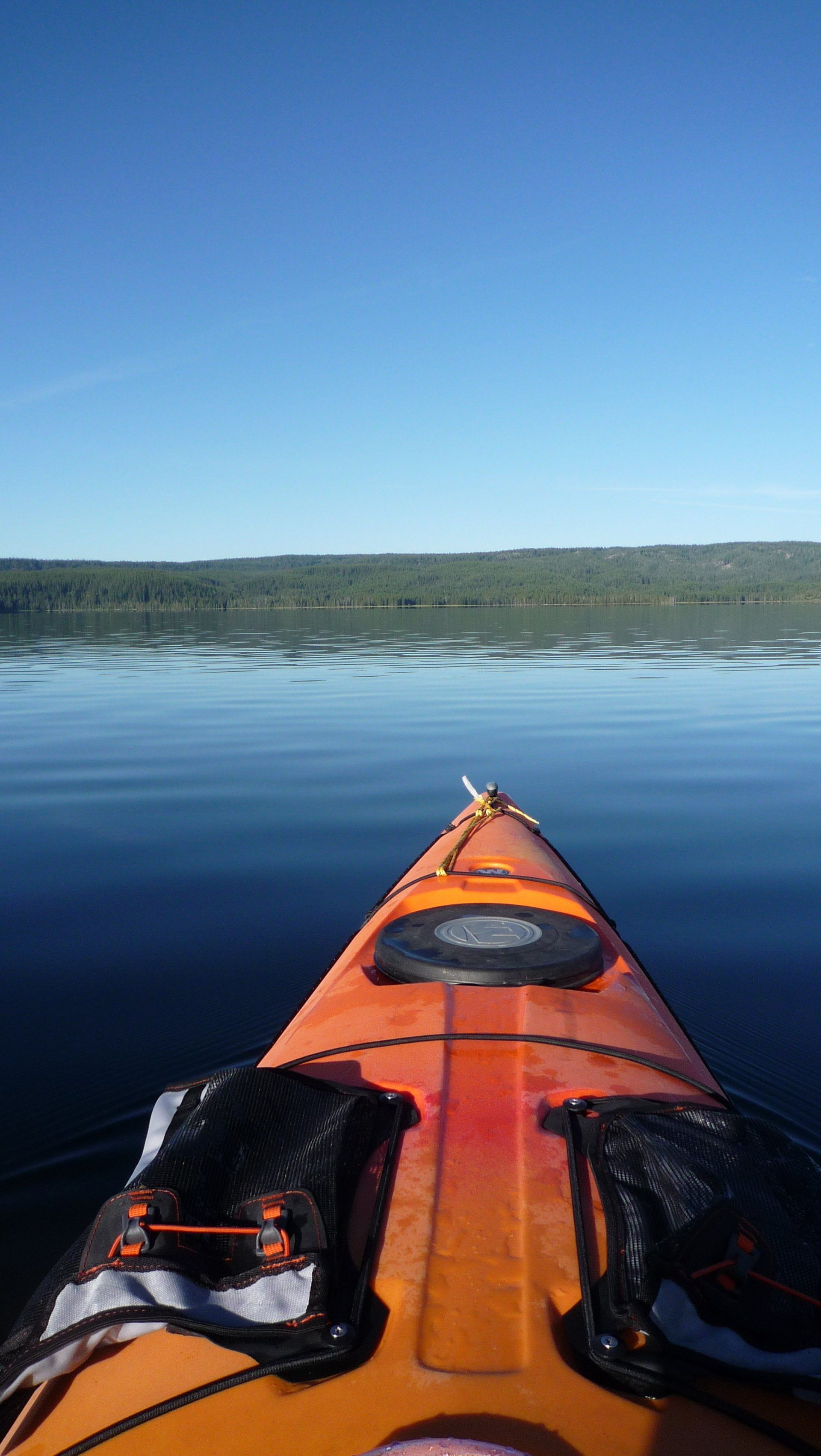 Orange kayak bow on calm blue water with distant tree-lined shore under clear sky