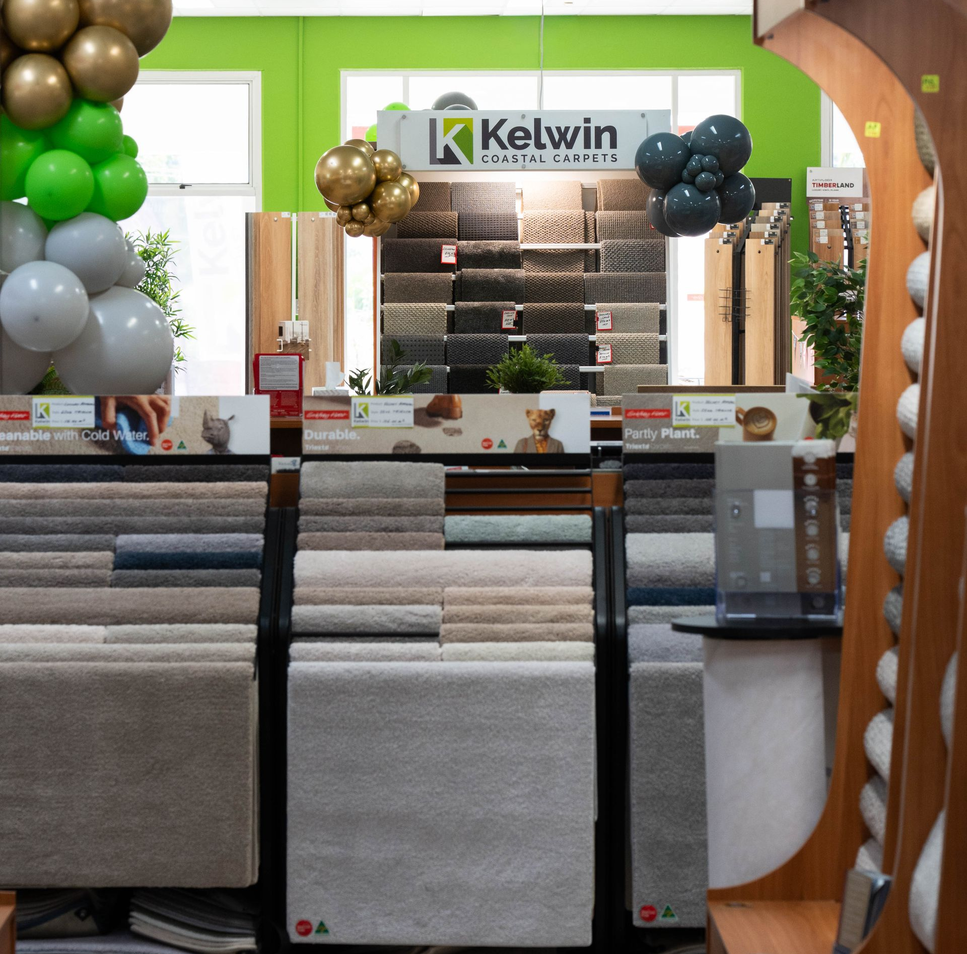 Carpet showroom with display racks, green walls, and balloon decorations around a Kelvin sign. — Kelwin Coastal Carpets In Kundra Park, QLD