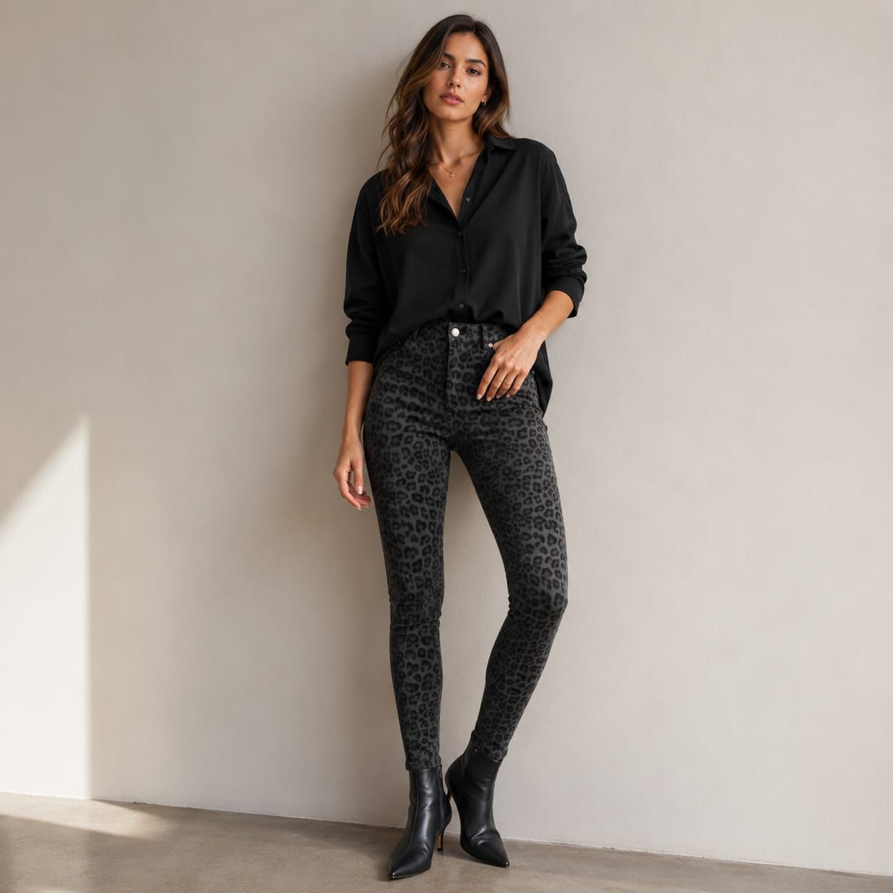 Mujer de pie apoyada contra una pared, con blusa negra, pantalones estampados y botines.