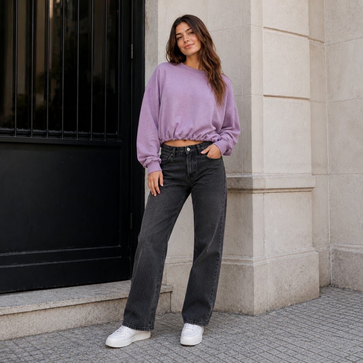 Mujer con suéter lila y jeans negros posando contra un muro de piedra junto a una puerta negra.