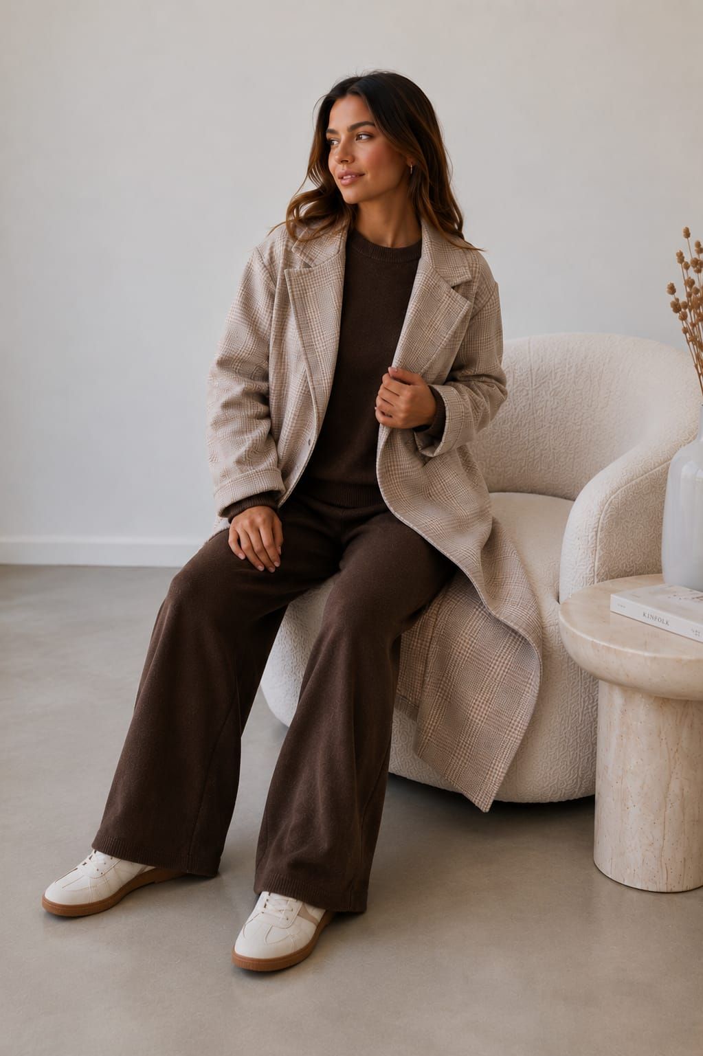 Persona sentada en una silla en una sala de estar de color beige, vestida con un abrigo a cuadros y pantalones marrones.