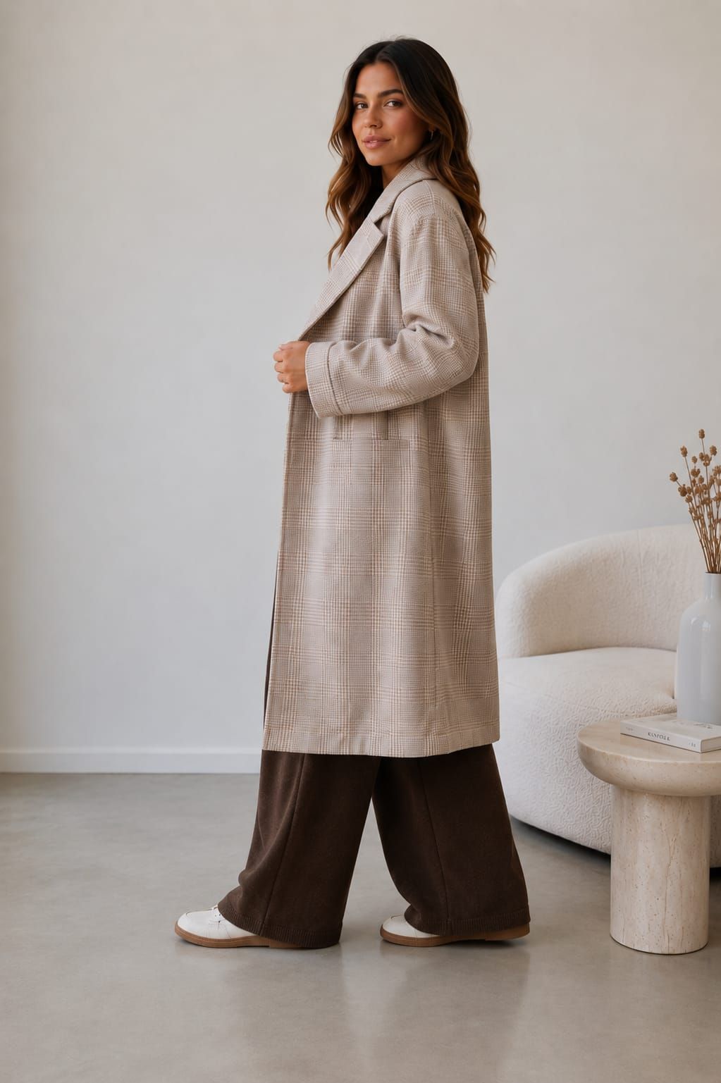 Persona sentada en una silla en una sala de estar de color beige, vestida con un abrigo a cuadros y pantalones marrones.