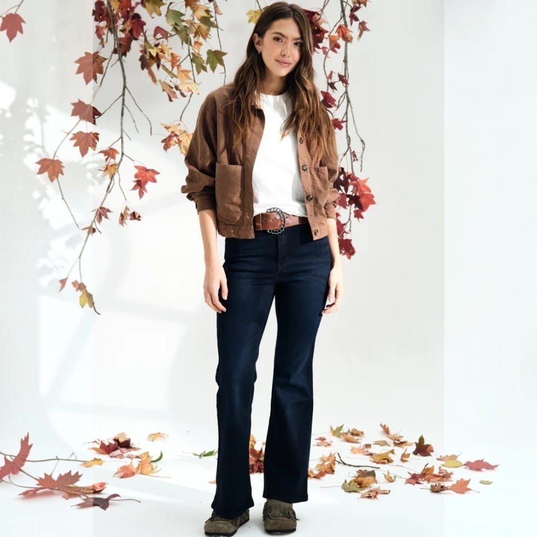 Mujer de pie en una habitación blanca con hojas de otoño, vestida con una chaqueta marrón y vaqueros oscuros.