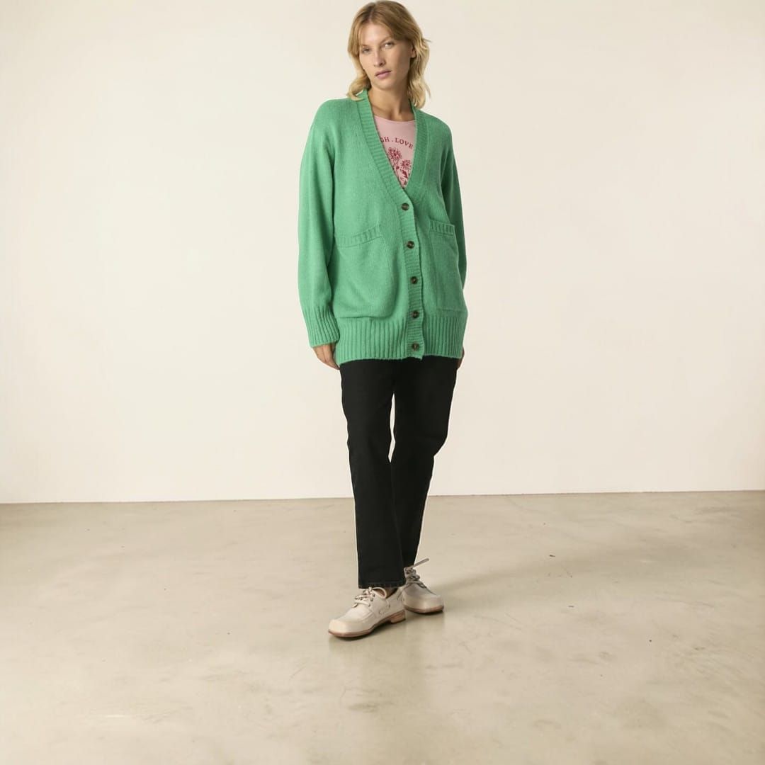 Persona de pie en un estudio con un cárdigan verde, una blusa rosa, pantalones negros y zapatillas blancas.