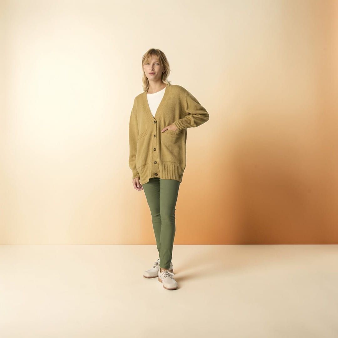 Persona de pie en un estudio con un cárdigan verde, una blusa rosa, pantalones negros y zapatillas blancas.
