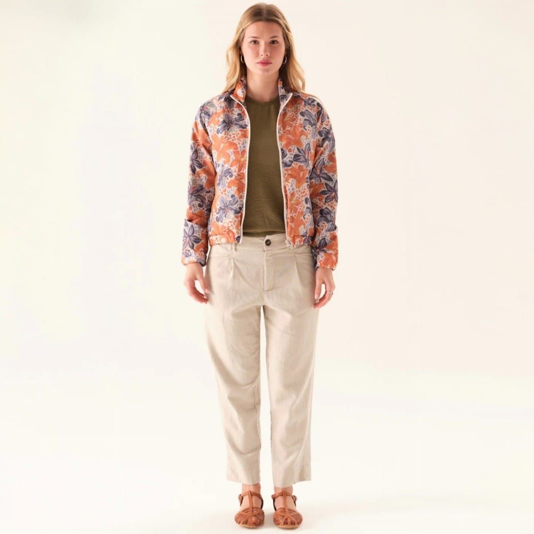 Mujer de pie con chaqueta floral, top marrón, pantalones beige y sandalias sobre un fondo blanco.