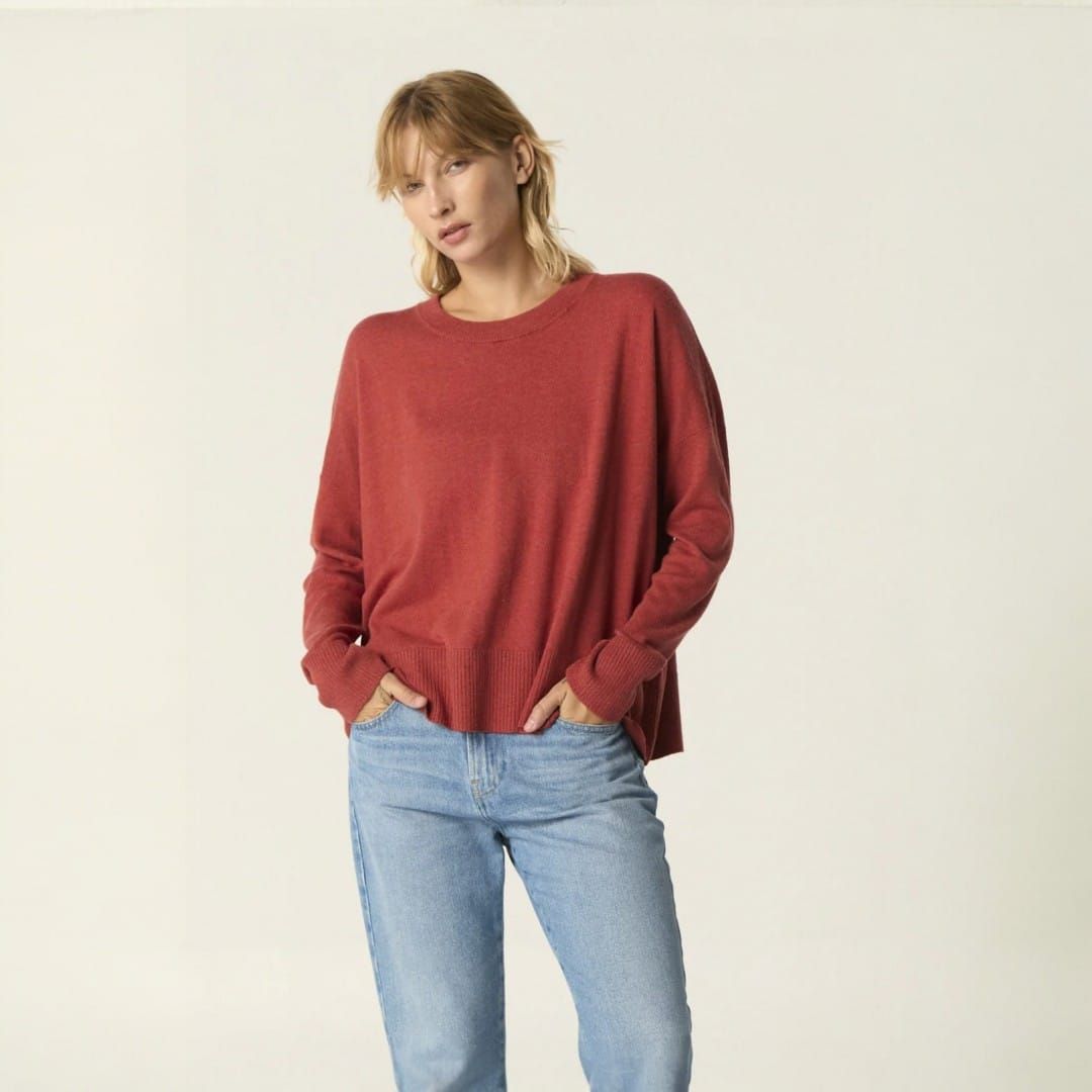 Persona con suéter rojo y jeans de pie sobre un fondo claro liso.