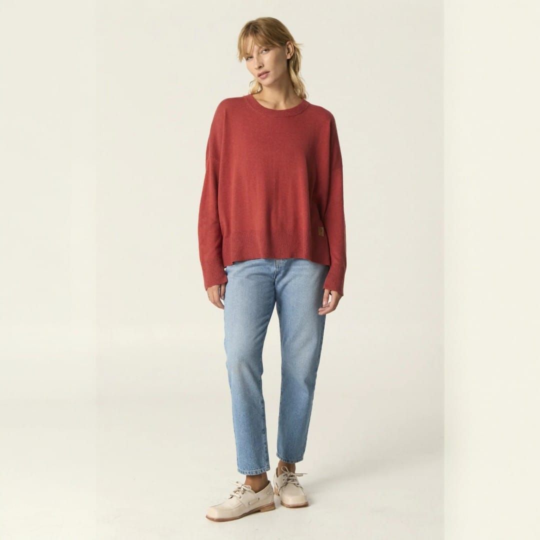 Persona con suéter rojo y jeans de pie sobre un fondo claro liso.