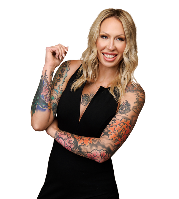 Woman with blonde hair and tattoos, wearing a black dress, smiles with crossed arms.