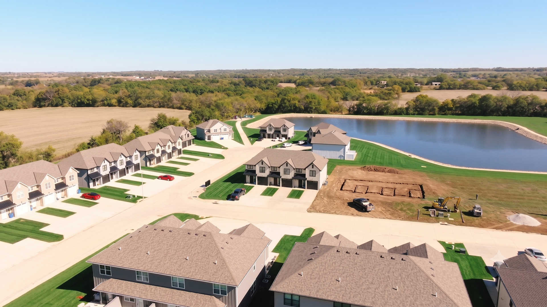 Aerial view of a suburban neighborhood with houses, a central road, and a lake beside green lawns