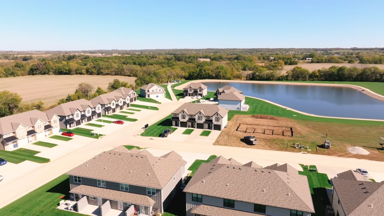 Aerial view of a suburban housing development beside a lake with townhomes, roads, and open land