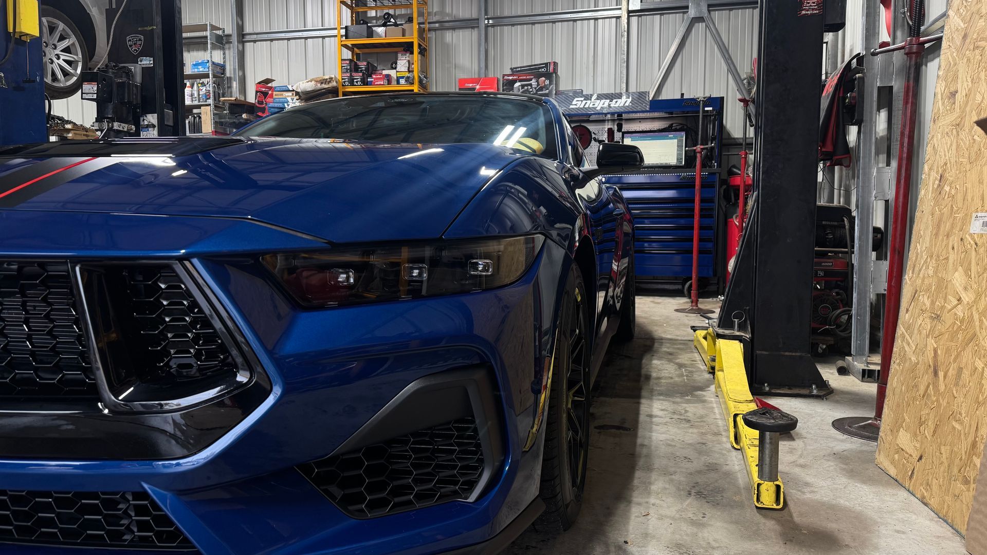 Blue Ford Mustang in a repair shop, black grill, red stripe, and bright yellow lift. | Alberto's Transmissions & Auto