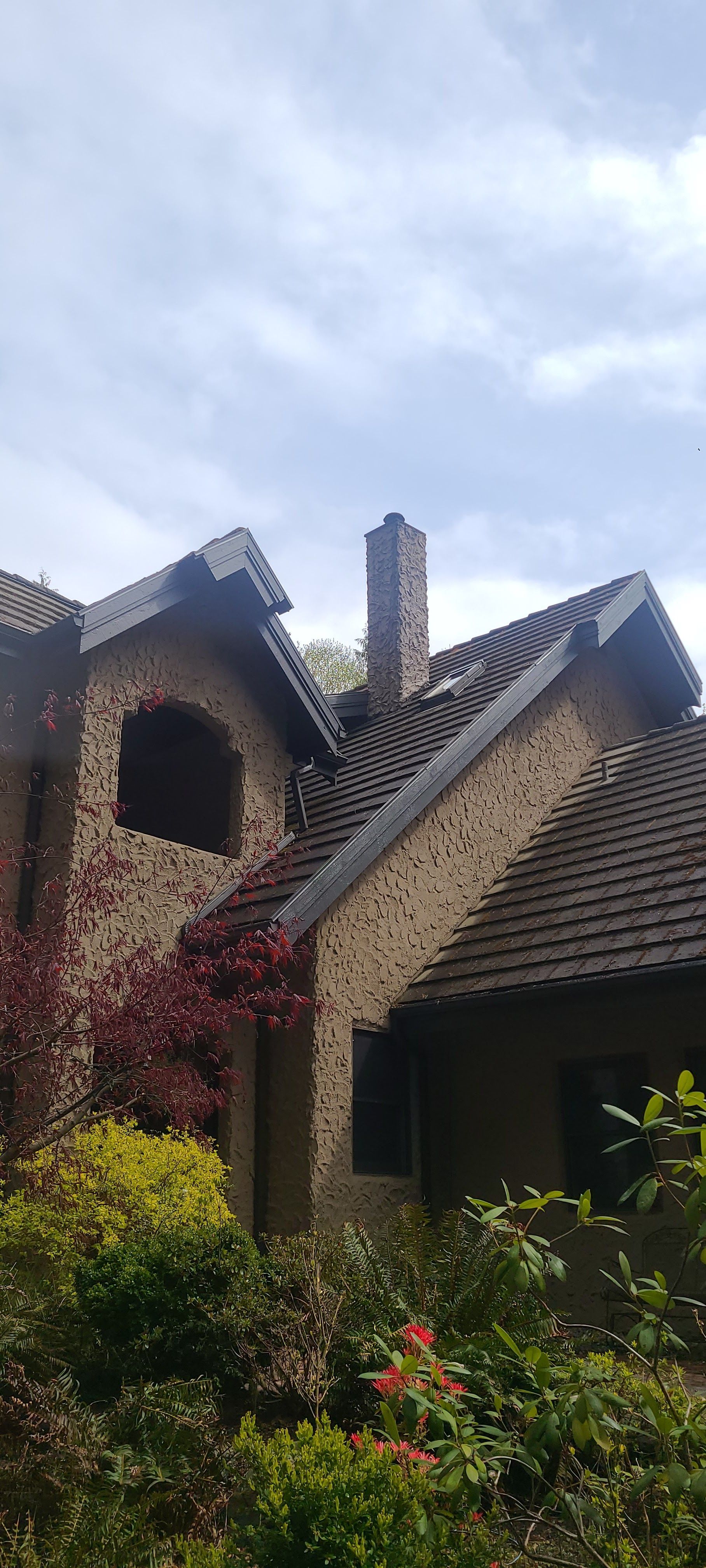 Brick house with steep gabled roof, chimney, and lush garden under a cloudy sky