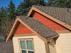House roof with brown shingles, beige siding, and red gable accents against trees and sky