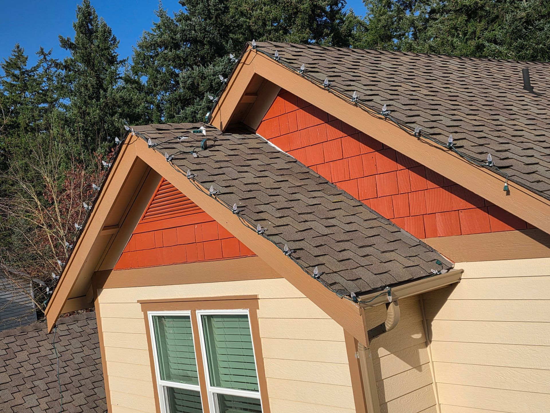 House roof with brown shingles, beige siding, and red gable accents against trees and sky