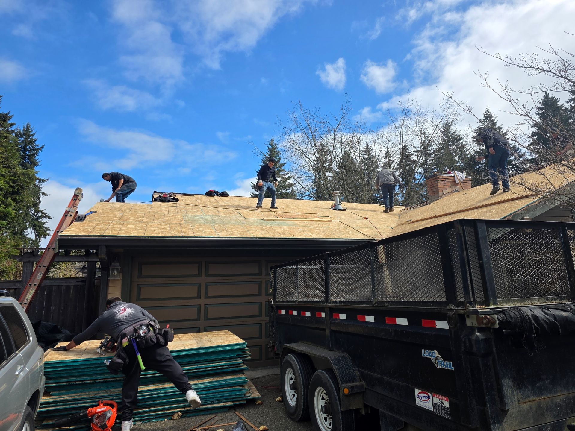Workers roofing a house, stacking shingles beside a black dump trailer under a blue sky.