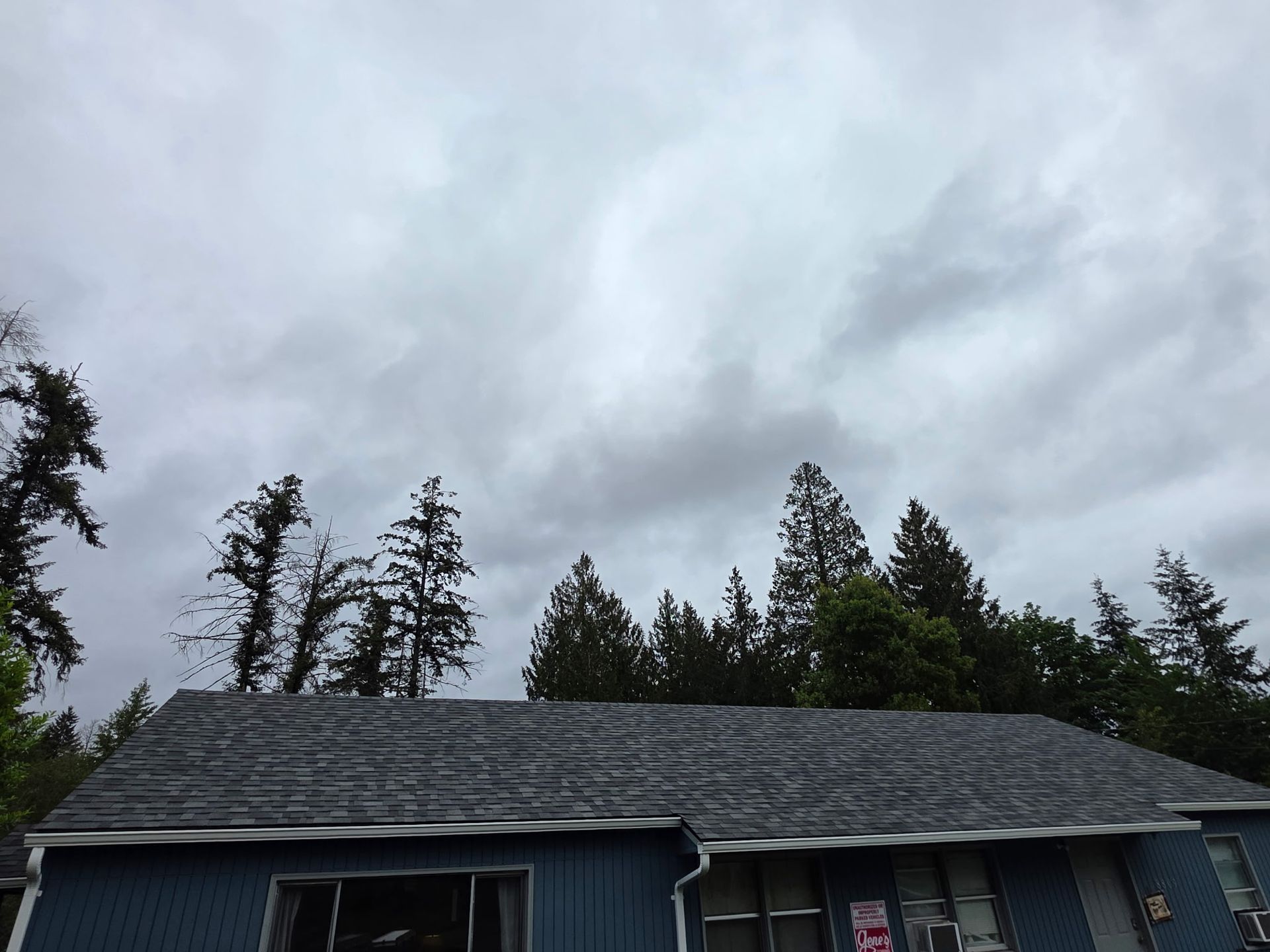 Cloudy sky over a house with dark roof and tall trees in the background.