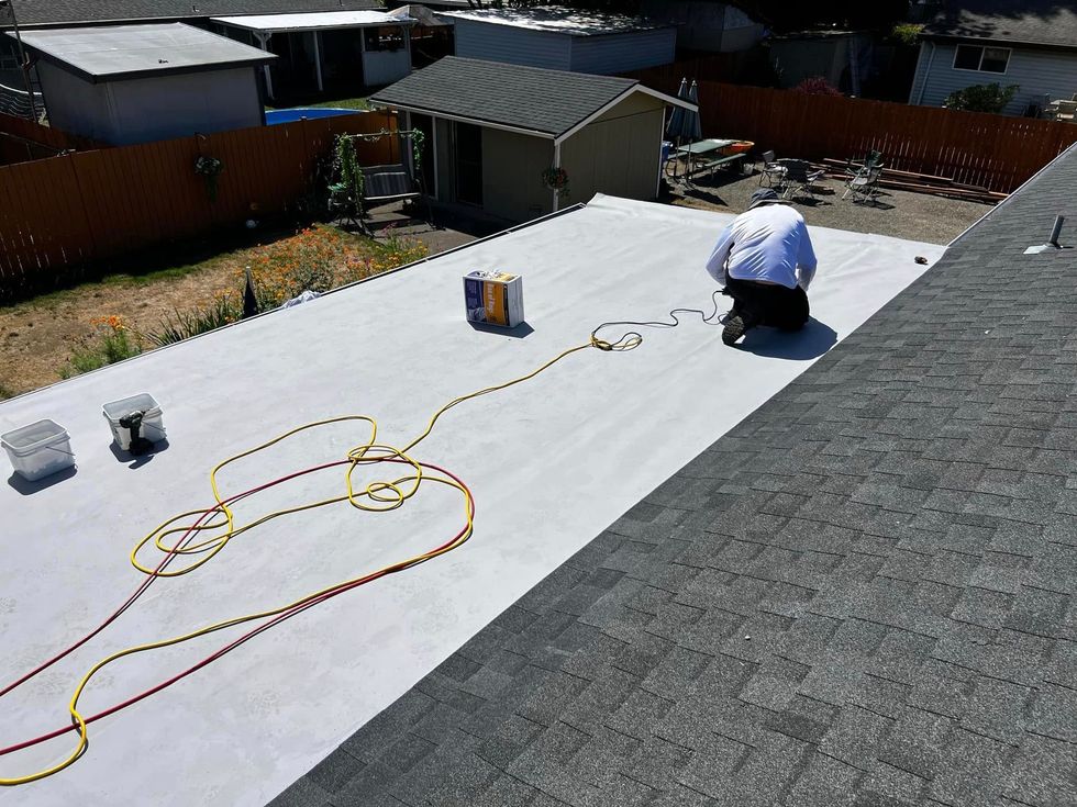 Worker kneeling on a white roof, inspecting materials and cords near the shingled edge.