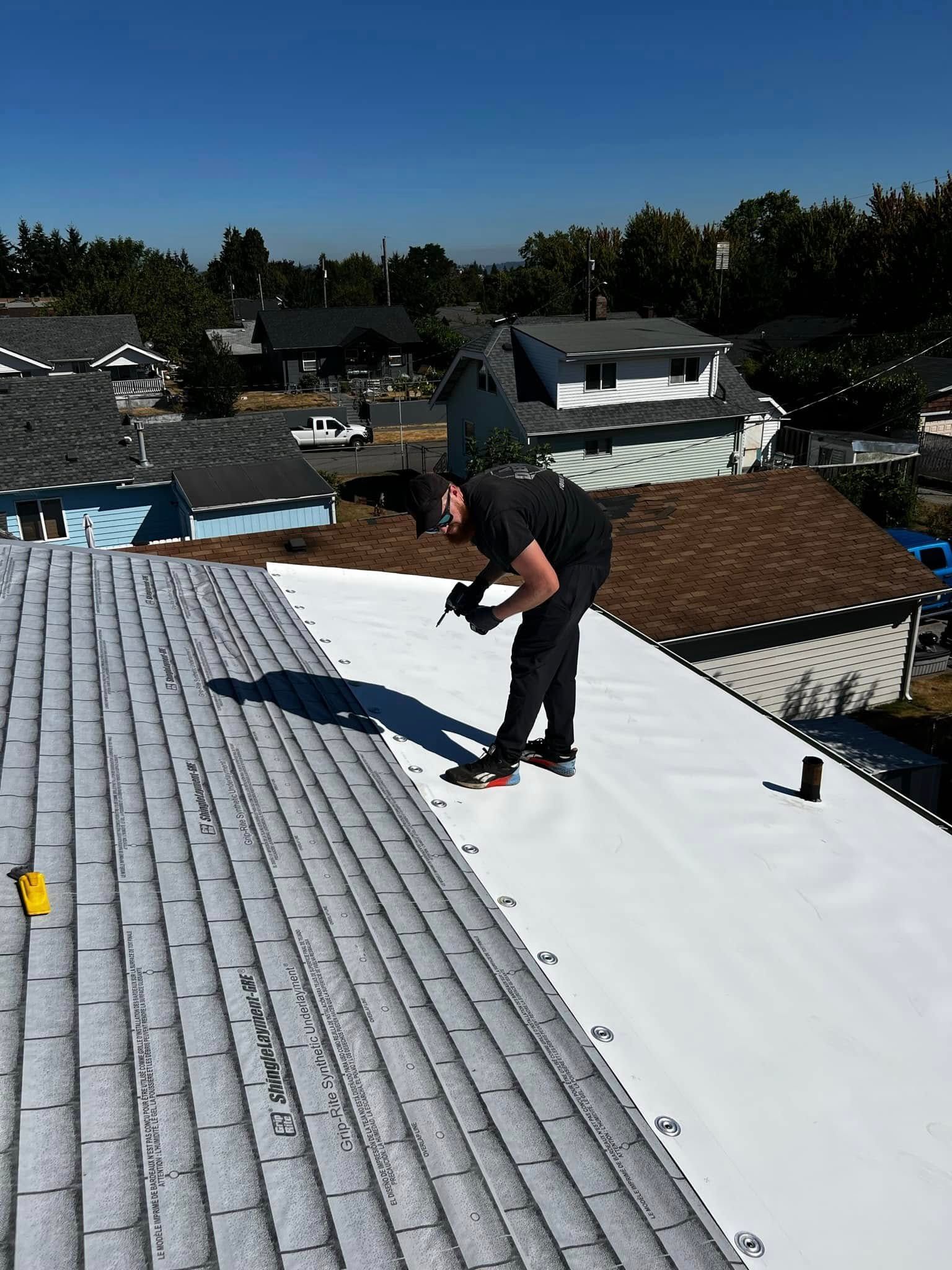 Worker on a rooftop beside gray shingles and white roof under clear sky.