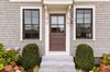 Front entrance of a house with gray siding, brown door, and two windows, framed by trimmed shrubs and flowers