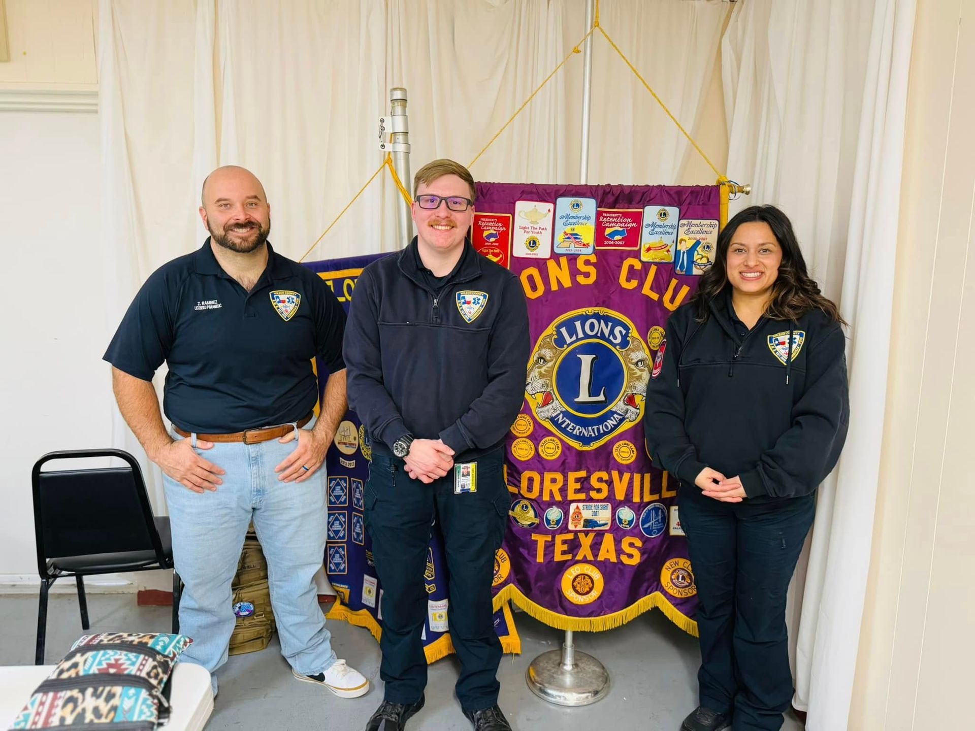 Three people are standing in front of a Lion 's club banner.