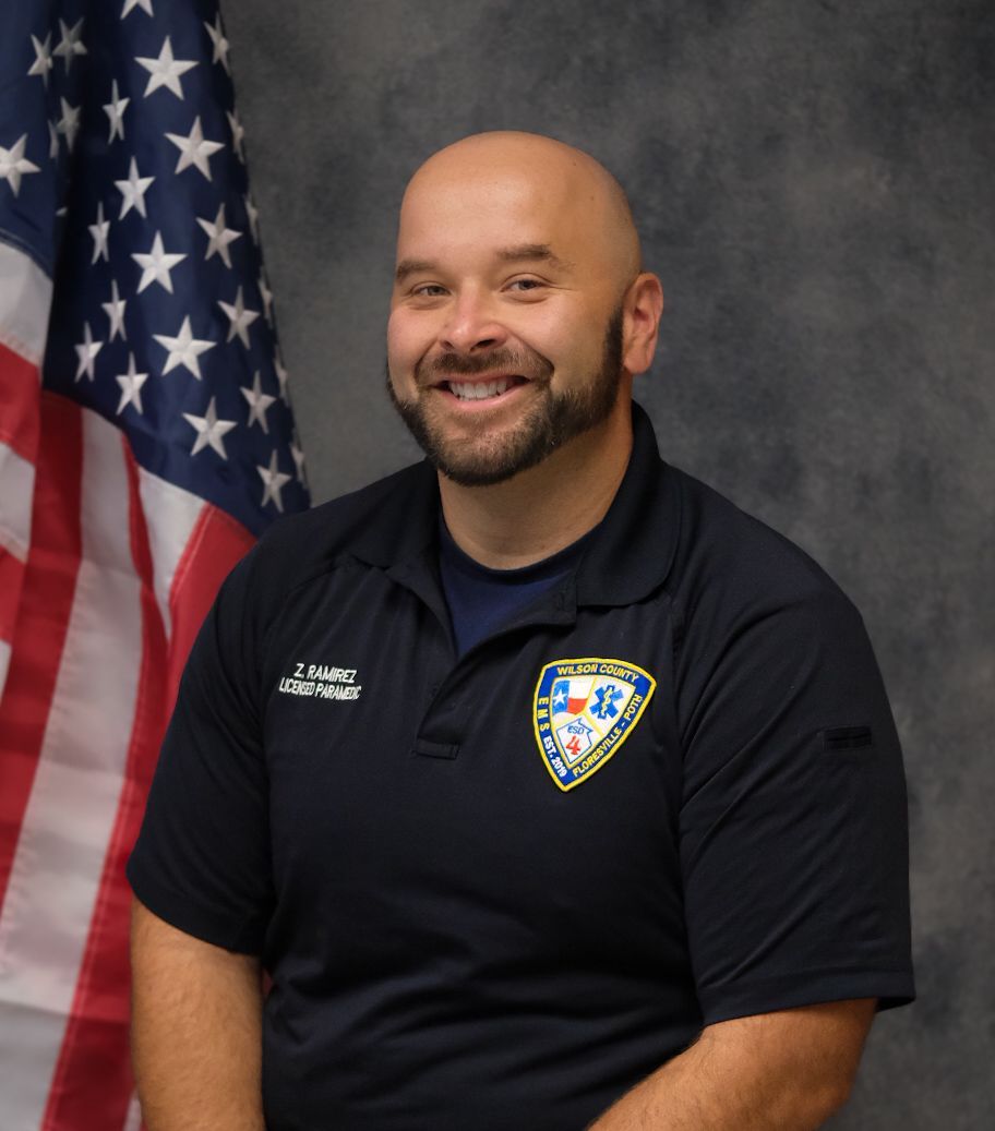 Smiling man in dark polo with EMS department patch, seated beside an American flag in a studio setting