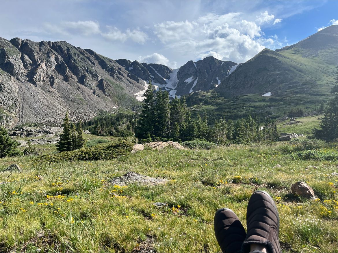 Mountains with patches of snow and green valley, person's feet in foreground, blue sky with clouds.