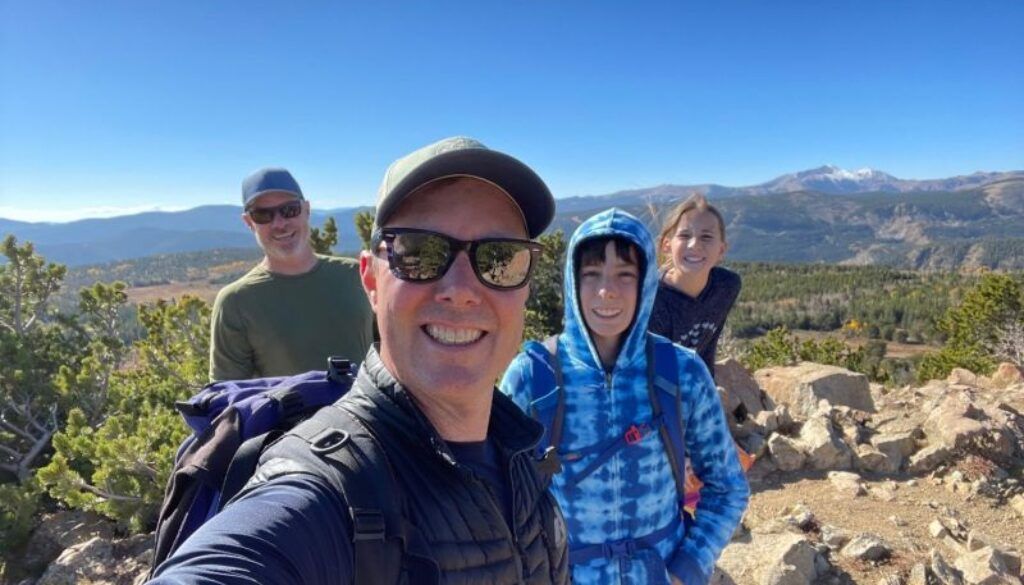 Three people rest on rocky mountaintop under blue sky, overlooking valley, wearing hats and backpacks.