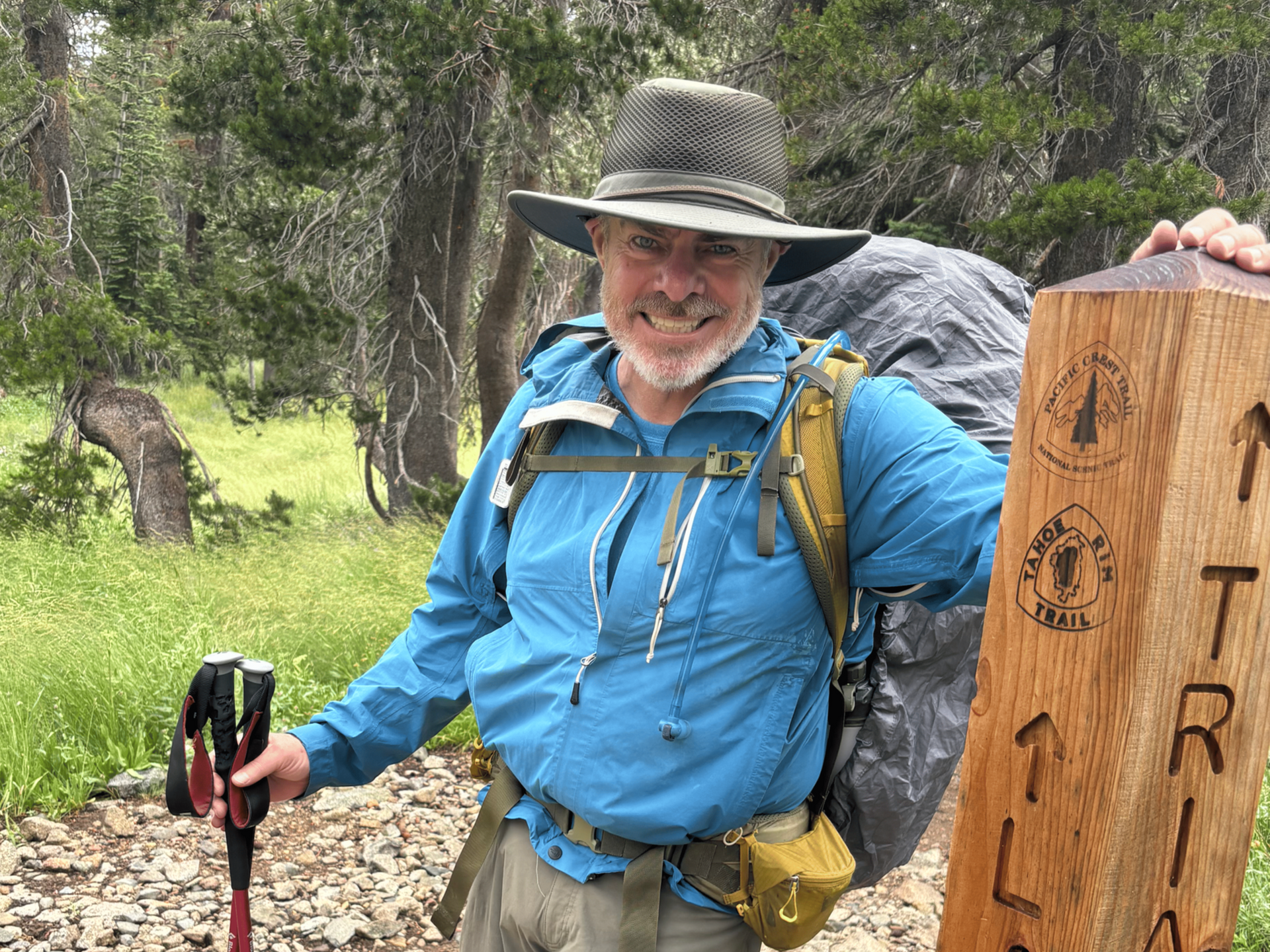 Man with hat and backpack smiles, standing next to a wooden trail marker in a wooded area.
