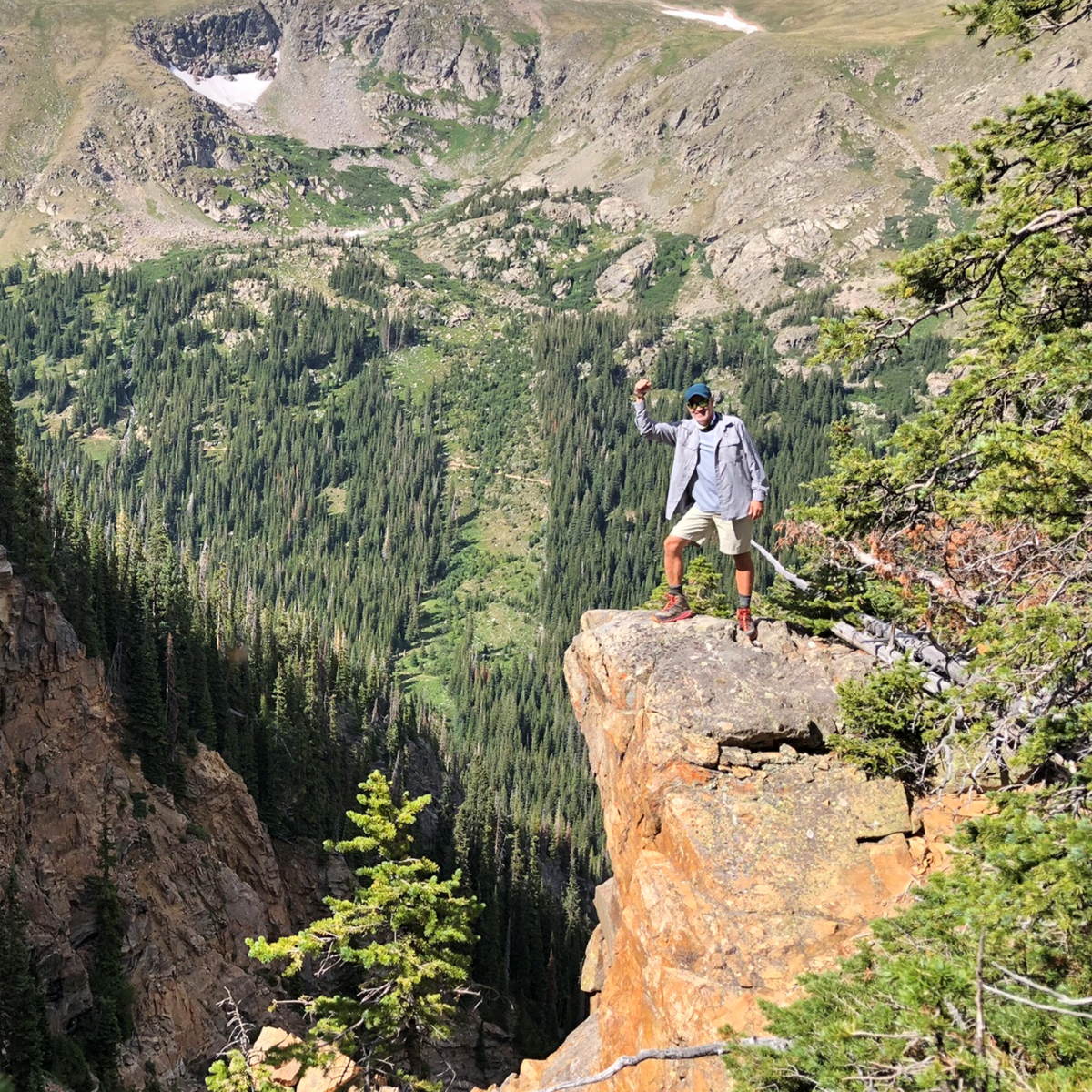 Man on cliff edge, arms raised, overlooking a deep, forested valley. Mountains in background.