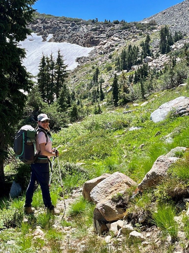 Person hiking on a grassy trail in a mountainous area, wearing a backpack and looking at the scenery.