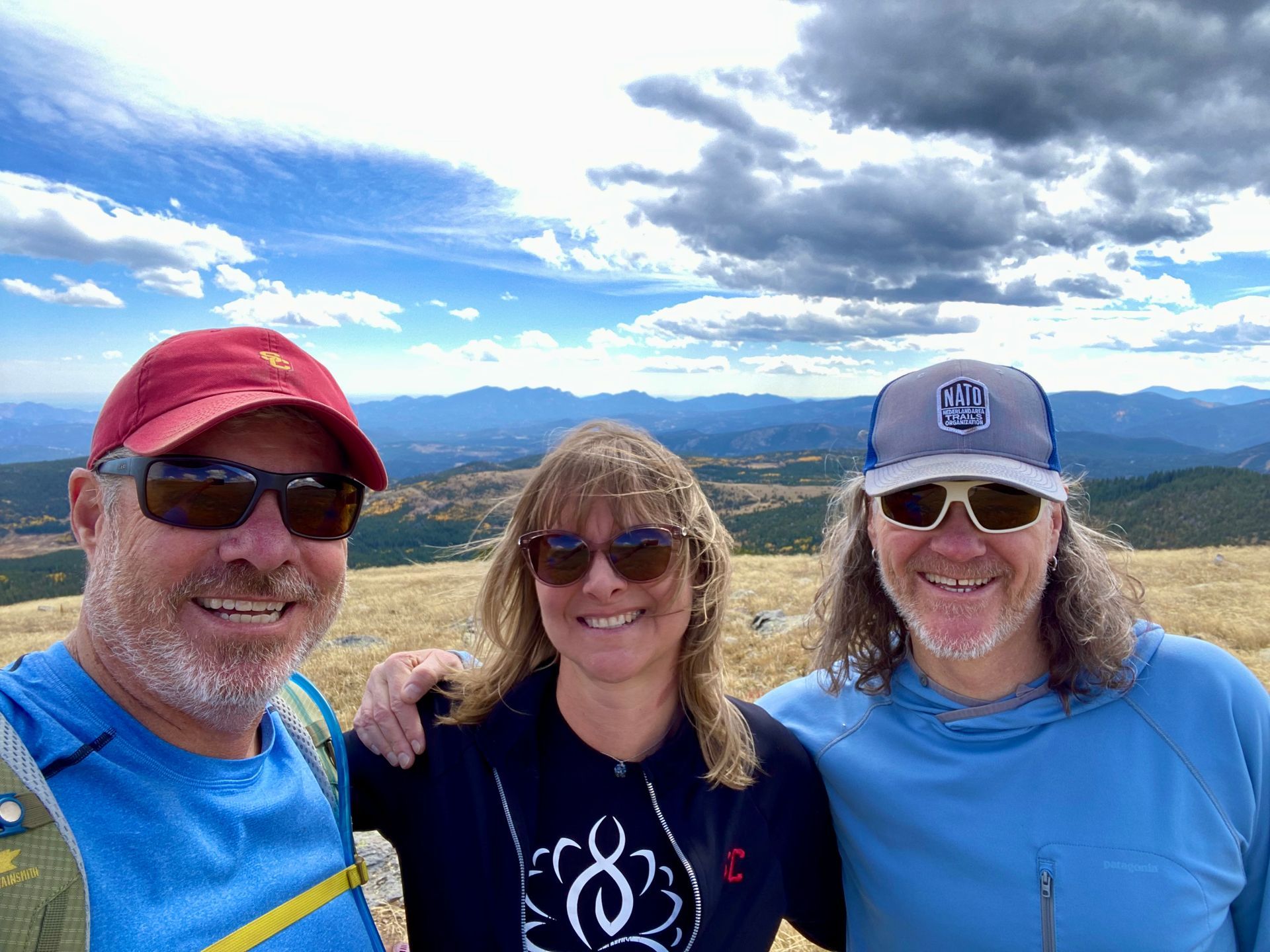 Three people smiling on a mountaintop with a cloudy sky. One man in a red cap, one woman, one man with a blue hoodie.